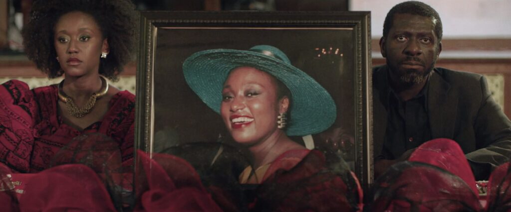 Sarah and Godwin sit at Grace's funeral with a portrait of Grace set between them